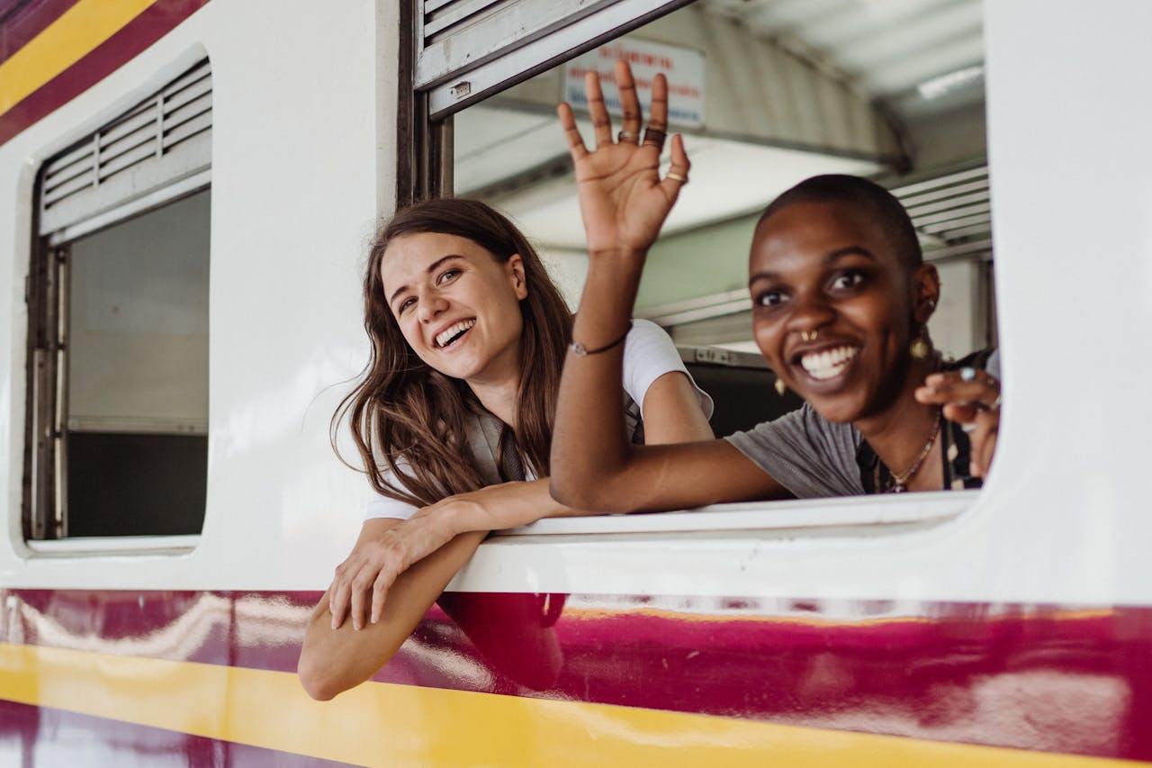 Two happy women on a train journey, smiling and waving through the window.